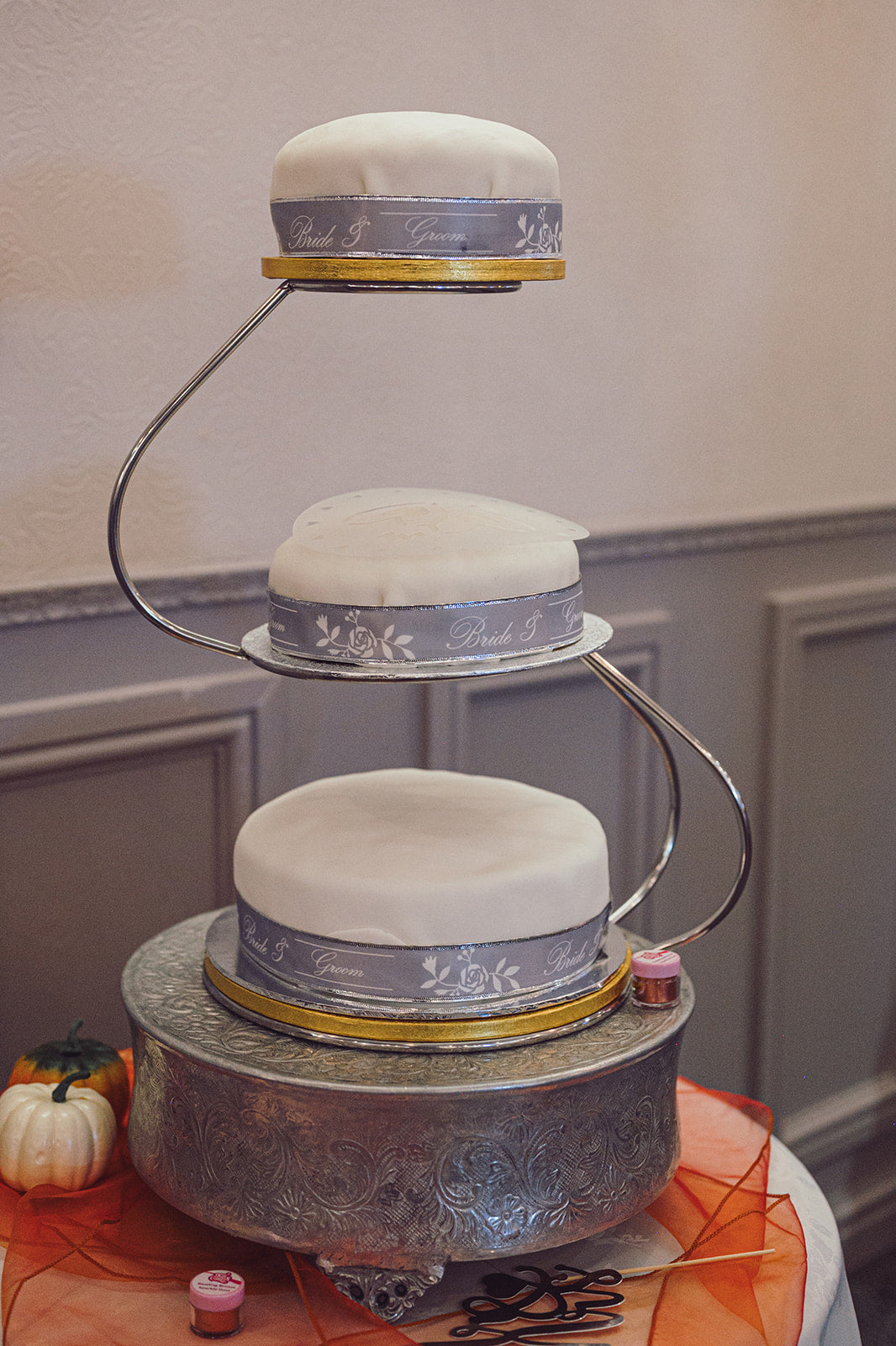 Three-tiered wedding cake with decorative ribbons on a stand against a neutral wall.